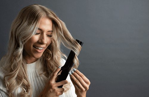 Woman styling blonde hair with a curling iron, smiling.